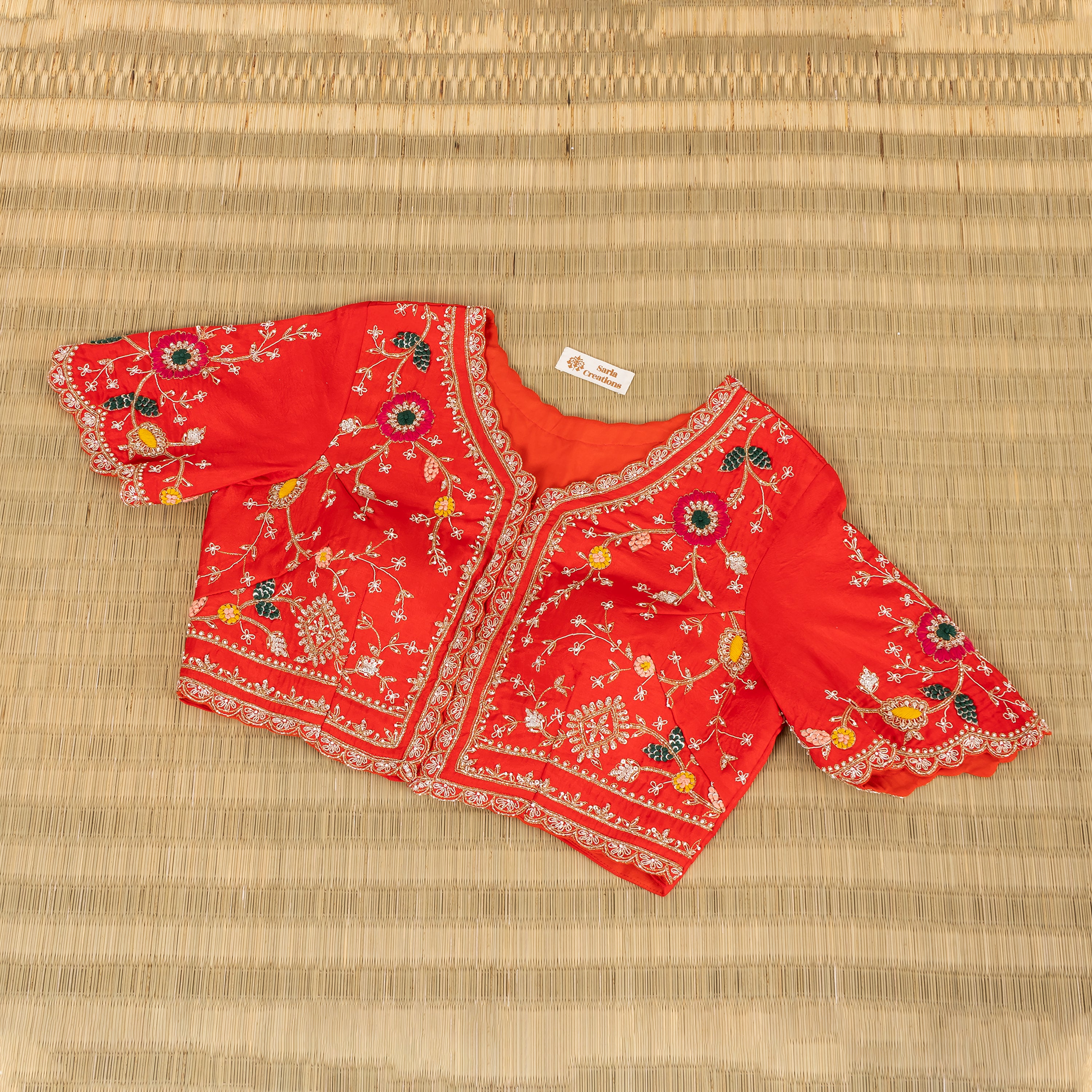 A red short-sleeve cropped blouse with intricate floral embroidery and scalloped borders, laid flat on a woven straw mat background.