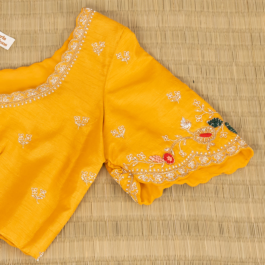 A yellow embroidered blouse top with short flared sleeves, laid flat on a woven straw mat. The blouse features intricate white embroidery, floral motifs in pink, red, and green, and a small white brand label inside the neckline.