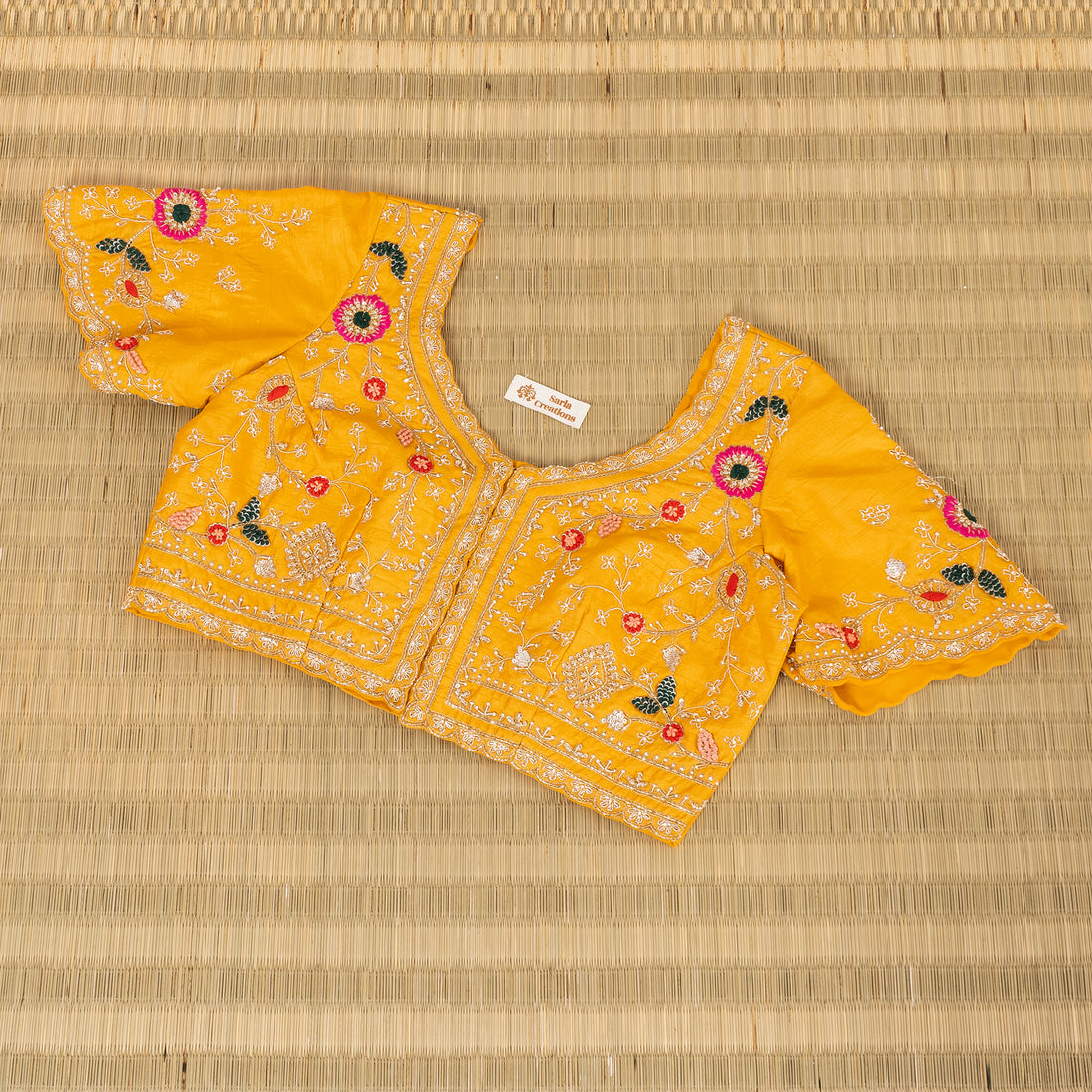 A yellow embroidered blouse top with short flared sleeves, laid flat on a woven straw mat. The blouse features intricate white embroidery, floral motifs in pink, red, and green, and a small white brand label inside the neckline.