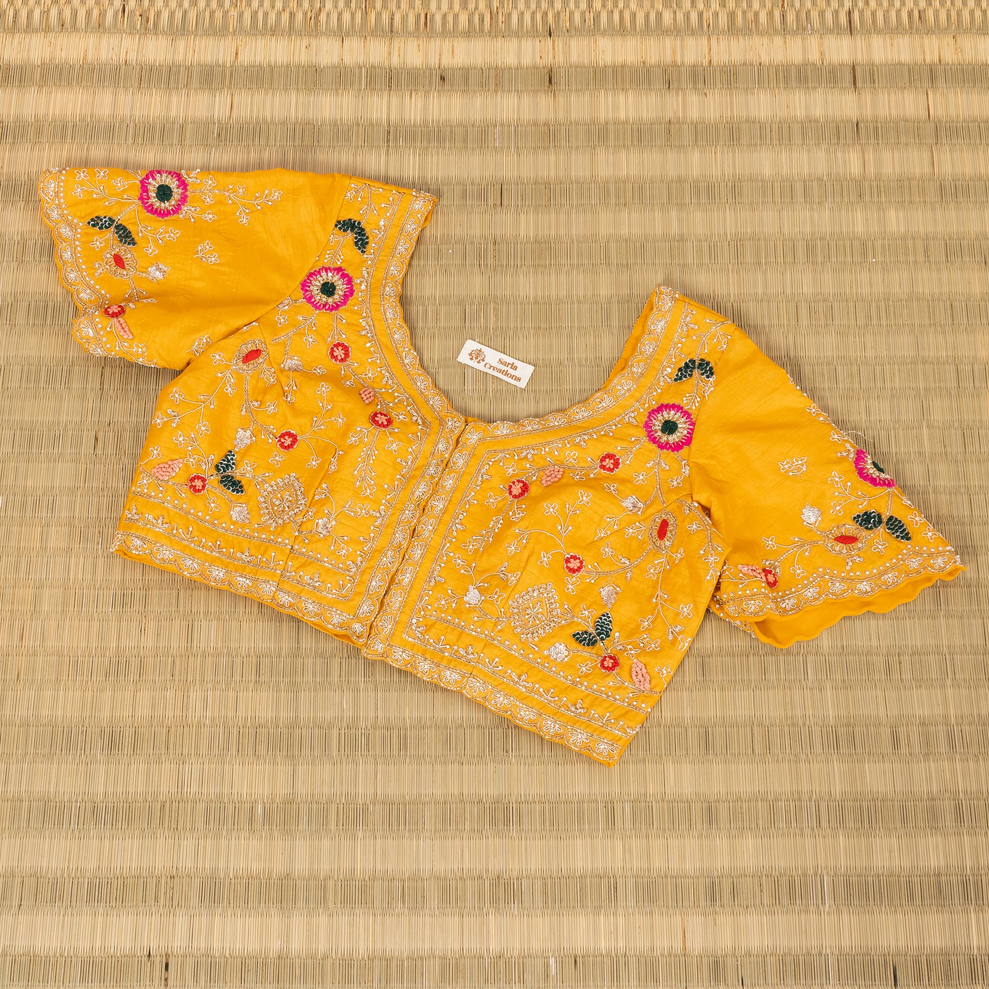 A yellow embroidered blouse top with short flared sleeves, laid flat on a woven straw mat. The blouse features intricate white embroidery, floral motifs in pink, red, and green, and a small white brand label inside the neckline.