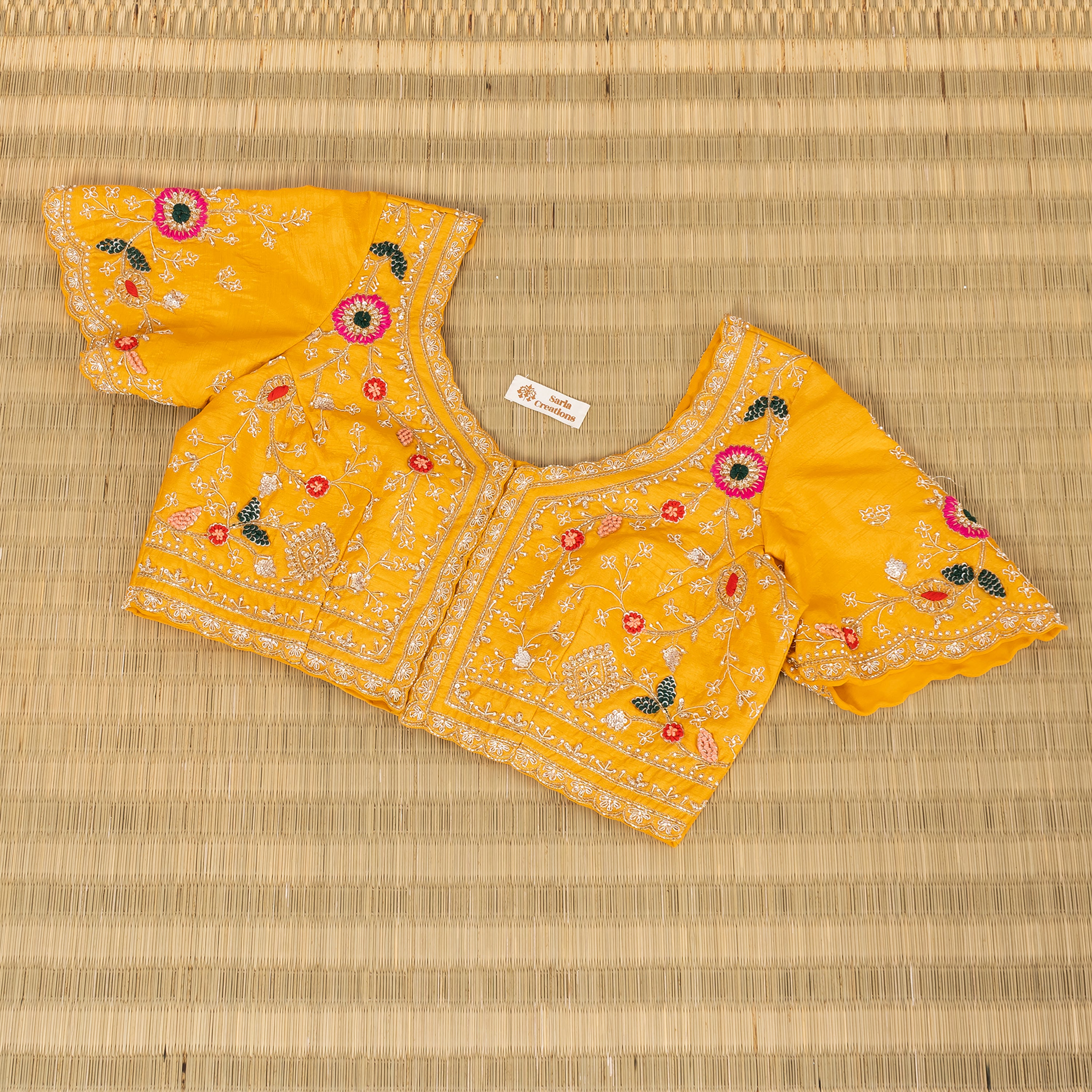 A yellow embroidered blouse top with short flared sleeves, laid flat on a woven straw mat. The blouse features intricate white embroidery, floral motifs in pink, red, and green, and a small white brand label inside the neckline.